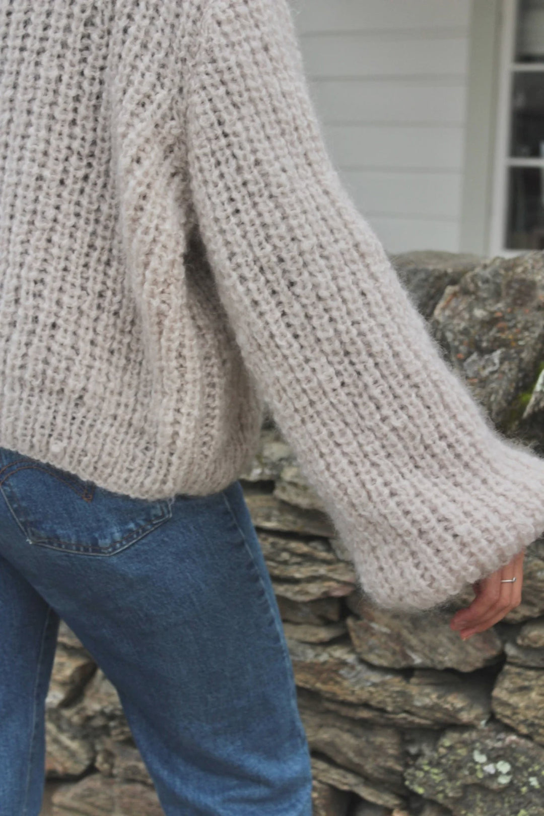Person wearing a chunky knit sweater and blue jeans standing against a stone wall.