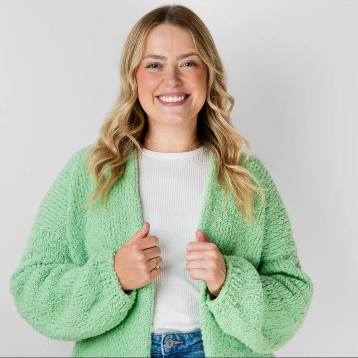 A smiling blonde woman wearing a light green fuzzy textured cardigan over a white ribbed top and blue jeans, posing against a light gray studio background.