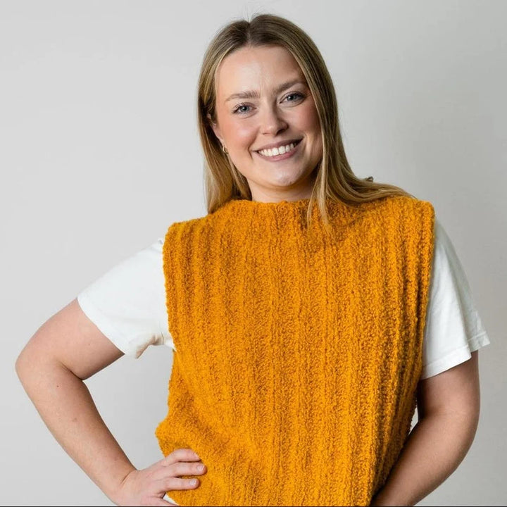 A smiling woman with blonde hair wearing a textured mustard yellow sleeveless knit top over a white t-shirt, posing against a light gray studio background.