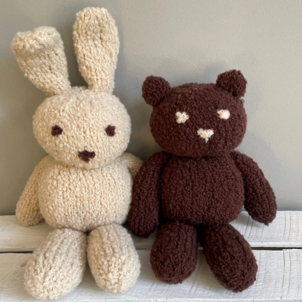 Two handmade, chunky-knit plush toys made from yarn: a cream-colored bunny with long ears and a dark brown teddy bear with cream-colored facial details, sitting side-by-side on a white wooden surface against a neutral gray wall.